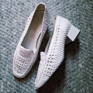 Pode incluir: Um par de sapatos brancos de couro tecido. Um é um mocassim slip-on e o outro é um sapato de salto baixo. Os sapatos apresentam um padrão de tecido de vime e estão sobre uma superfície texturizada azul-esverdeada clara.