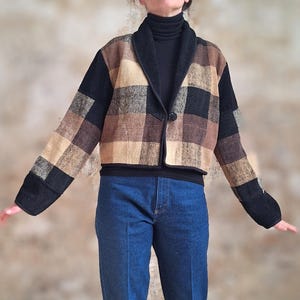 May include: A cropped jacket with a plaid pattern in shades of brown, tan, and black. The jacket has a black collar and cuffs, and a single button closure. The model is wearing blue jeans and a black turtleneck.