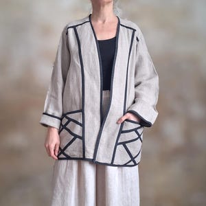 May include: A beige linen jacket with black trim and geometric pocket designs. The jacket has a relaxed fit and three-quarter sleeves. The model is wearing a black top and beige pants.