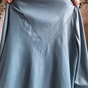 May include: A light blue leather coat with a V-shaped design on the back. The coat has long sleeves and a collar. The stitching is visible along the seams and the V-shaped design.