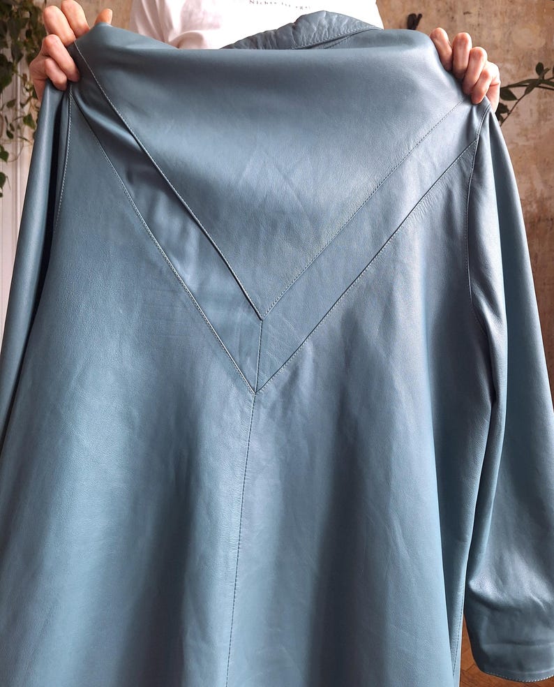 May include: A light blue leather coat with a V-shaped design on the back. The coat has long sleeves and a collar. The stitching is visible along the seams and the V-shaped design.