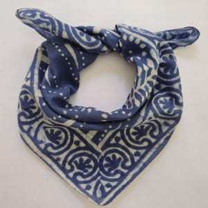 May include: A square, indigo blue bandana with a cream-colored, ornate pattern. The design features repeating floral and geometric shapes. The bandana is knotted at the top, showcasing the intricate print and soft fabric.
