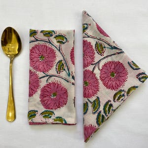 Op de afbeelding: Een gouden lepel ligt naast twee stoffen servetten. De servetten hebben een bloemenpatroon met roze bloemen, groene bladeren en blauwgroene stelen op een witte achtergrond. Eén servet is gevouwen, de andere plat.