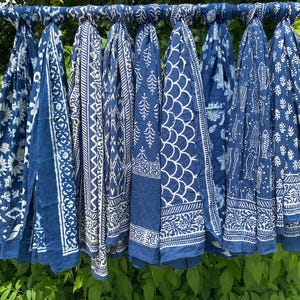Könnte beinhalten: Eine Sammlung von acht rechteckigen Stoffschals, die nebeneinander hängen und jeweils ein eigenes weißes Muster auf dunkelblauem Grund aufweisen. Die Designs umfassen florale Motive, geometrische Formen, Fische und geschwungene Muster.
