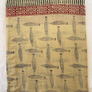 May include: A folded beige fabric with a repeating pattern of black fish. The fabric has a decorative border with a red geometric design and black stripes. The fish are stylized with vertical lines and fins.