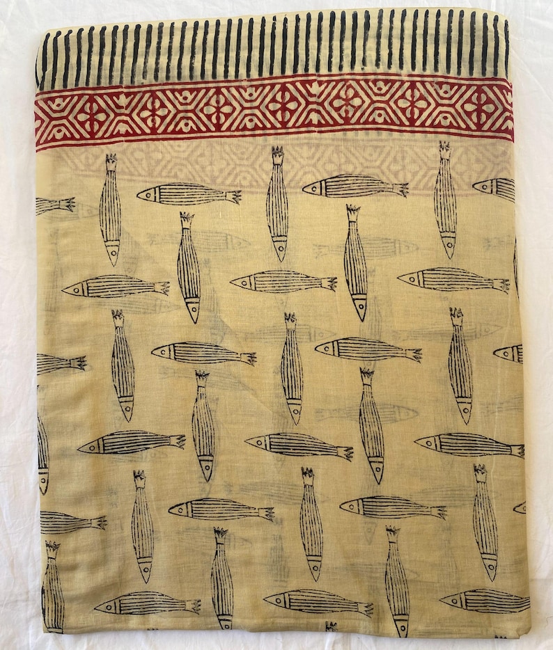 May include: A folded beige fabric with a repeating pattern of black fish. The fabric has a decorative border with a red geometric design and black stripes. The fish are stylized with vertical lines and fins.