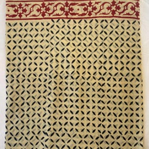 May include: A folded textile with a cream-colored background and a repeating pattern of black, oval-shaped designs. The top edge features a decorative border in red with floral motifs. The fabric appears to be lightweight and suitable for various uses.