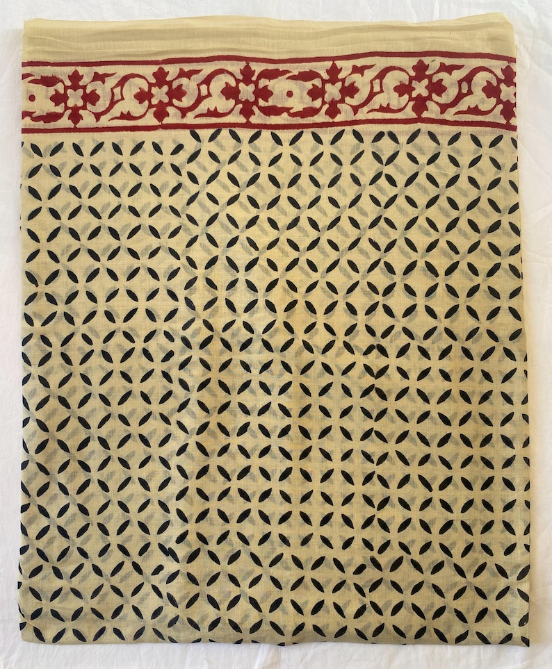 May include: A folded textile with a cream-colored background and a repeating pattern of black, oval-shaped designs. The top edge features a decorative border in red with floral motifs. The fabric appears to be lightweight and suitable for various uses.