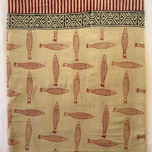 May include: Cream-colored fabric with a repeating pattern of red fish. The top border features a design of vertical red stripes and a geometric pattern. The fabric is likely cotton or linen, suitable for clothing or home decor.