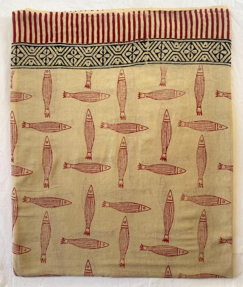 May include: Cream-colored fabric with a repeating pattern of red fish. The top border features a design of vertical red stripes and a geometric pattern. The fabric is likely cotton or linen, suitable for clothing or home decor.
