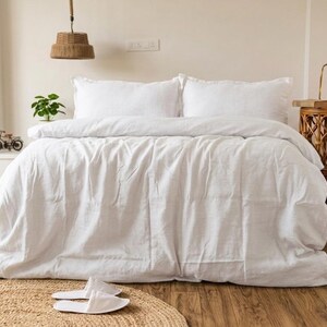 May include: A bed with white bedding, including a duvet and two pillows. A woven rug and white slippers are in the foreground. A decorative lamp and side table are on the right. A small plant and a decorative bicycle are on the left.