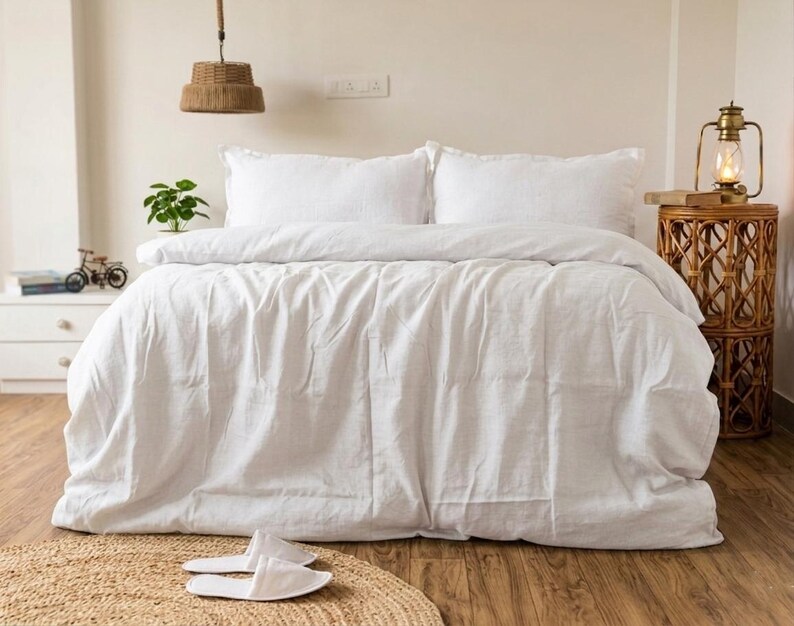 May include: A bed with white bedding, including a duvet and two pillows. A woven rug and white slippers are in the foreground. A decorative lamp and side table are on the right. A small plant and a decorative bicycle are on the left.