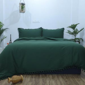 Könnte beinhalten: Ein dunkelgrünes Bettwäsche-Set mit zwei Kissenbezügen und einer Bettdecke, alle mit kleinen Pompons verziert. Das Bett ist gemacht und einsatzbereit. Der Raum ist minimalistisch gestaltet mit Pflanzen und einer dekorativen Laterne.