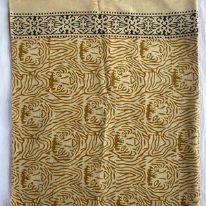 May include: A beige textile with a repeating pattern of stylized tiger faces in shades of brown. The top edge features a decorative black border with floral and dot motifs. The fabric appears to be cotton or linen.