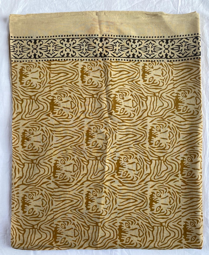 May include: A beige textile with a repeating pattern of stylized tiger faces in shades of brown. The top edge features a decorative black border with floral and dot motifs. The fabric appears to be cotton or linen.