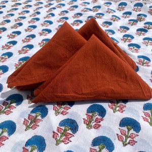 Puede incluir: Tres servilletas de algodón de color óxido dobladas en forma de triángulo, dispuestas sobre un fondo de tela blanca con un patrón floral azul y rosa.
