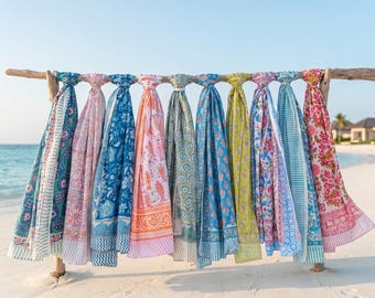 May include: A collection of colorful, patterned scarves hanging on a wooden rack at the beach. The scarves feature various designs, including floral, paisley, and geometric patterns, in shades of blue, pink, green, and yellow. The scarves are made of lightweight fabric.