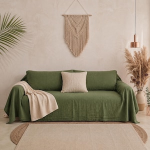 May include: A sage green linen sofa cover draped over a couch, featuring a textured surface. A cream-coloured square throw pillow rests in the centre, and a folded beige knit throw blanket is placed over the left arm of the sofa.