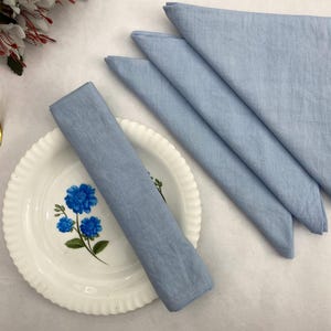 Puede incluir: Una naturaleza muerta de una mesa con un tenedor dorado, un plato blanco con un diseño floral azul y servilletas azul claro dobladas. Las servilletas están dispuestas en forma de abanico, y una está enrollada. Flores decorativas en el fondo.
