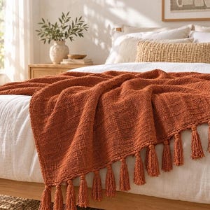 May include: A textured, burnt orange throw blanket featuring a woven grid pattern and a row of matching fringe tassels along the bottom edge, draped over a white bedspread with neutral-toned pillows in the background.