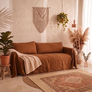 May include: A rust-colored linen sofa cover draped over a couch, featuring a textured cream-colored throw blanket. The sofa is positioned in a room with a macrame wall hanging, a hanging potted plant, a fiddle leaf fig, and a patterned rug.