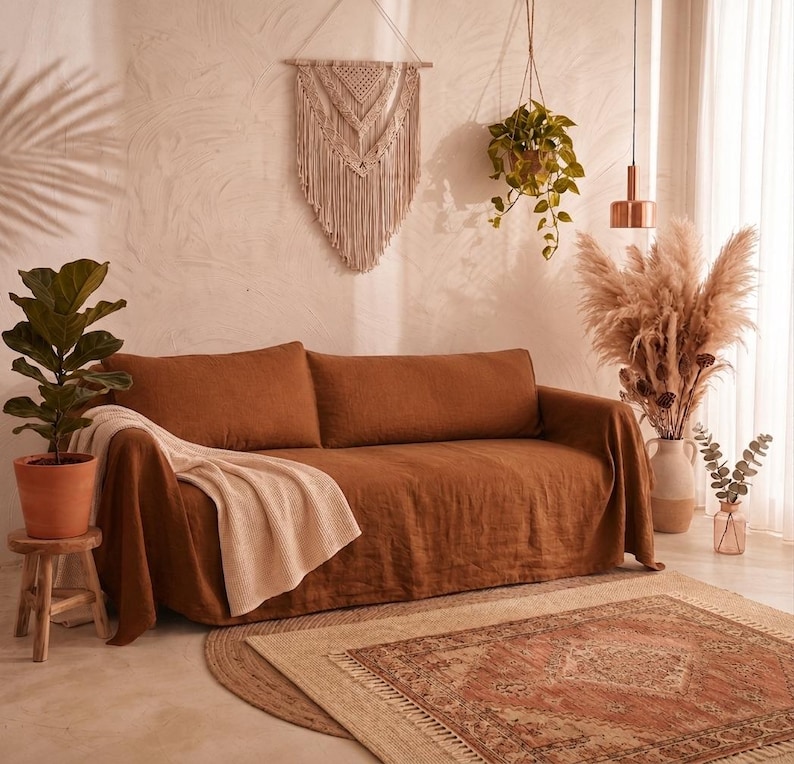 May include: A rust-colored linen sofa cover draped over a couch, featuring a textured cream-colored throw blanket. The sofa is positioned in a room with a macrame wall hanging, a hanging potted plant, a fiddle leaf fig, and a patterned rug.