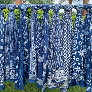 Puede incluir: Una colección de bufandas azul índigo con patrones blancos, colgadas de una barra de metal. Las bufandas muestran varios diseños, incluyendo motivos florales, geométricos y paisley. La tela es ligera, y las bufandas se exhiben al aire libre.