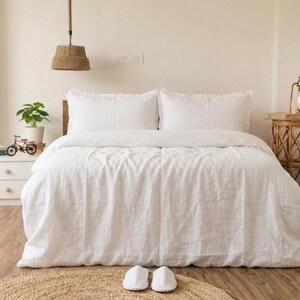 May include: A bedroom scene featuring a bed with white bedding, including a duvet and pillows. A small side table with a lamp and a decorative woven basket are on the right. A white nightstand with a plant and a miniature bicycle is on the left.