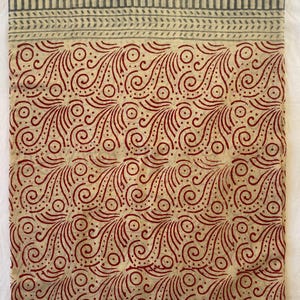 May include: A folded textile with a cream-colored base and a repeating pattern of red swirls and dots. The top border features a striped design in gray. The fabric is likely cotton or linen, suitable for clothing or home decor.
