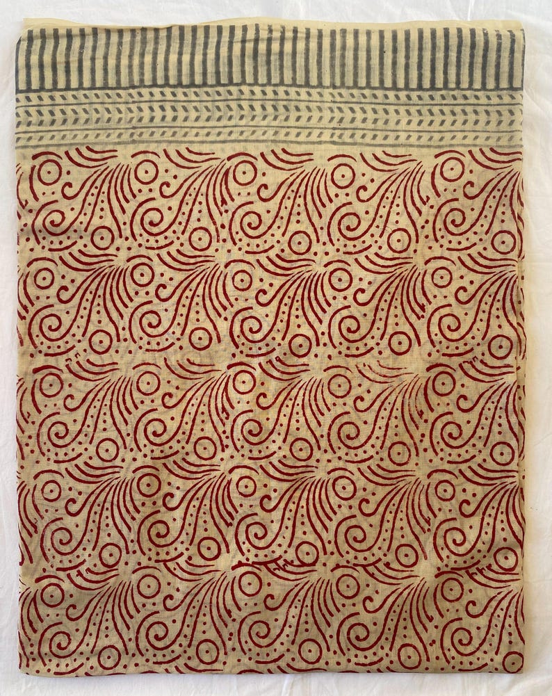 May include: A folded textile with a cream-colored base and a repeating pattern of red swirls and dots. The top border features a striped design in gray. The fabric is likely cotton or linen, suitable for clothing or home decor.