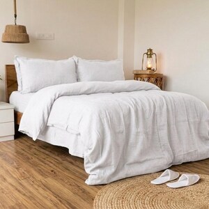 May include: A bed with white bedding, including a duvet and pillows, is the focal point of this bedroom. The bed sits on a wooden frame, and the room features a wooden floor and a woven lamp hanging from the ceiling. Two white slippers rest on a round rug.