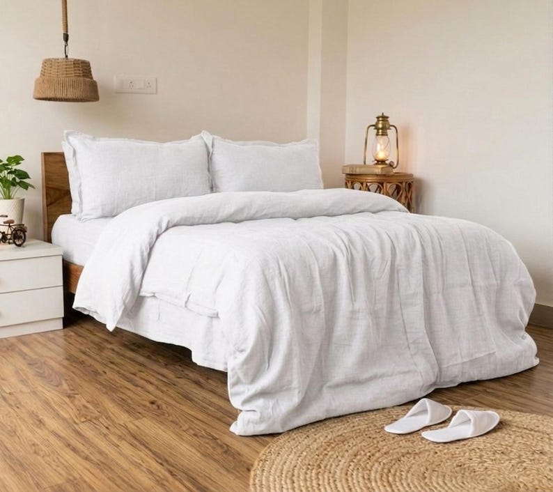 May include: A bed with white bedding, including a duvet and pillows, is the focal point of this bedroom. The bed sits on a wooden frame, and the room features a wooden floor and a woven lamp hanging from the ceiling. Two white slippers rest on a round rug.
