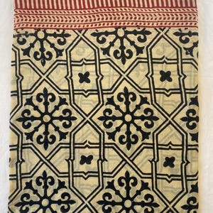 May include: Folded fabric with a cream-colored background and a repeating black geometric pattern. The top edge features a red border with vertical stripes and a decorative band. The fabric is likely used for home decor.