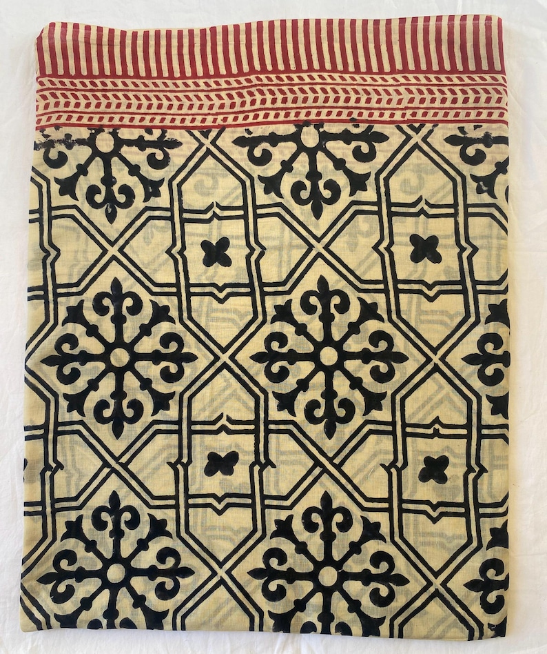May include: Folded fabric with a cream-colored background and a repeating black geometric pattern. The top edge features a red border with vertical stripes and a decorative band. The fabric is likely used for home decor.