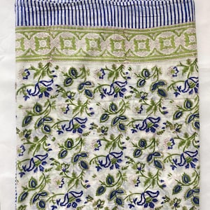 May include: A folded white fabric with a blue and green floral pattern. The top edge features blue and white vertical stripes, followed by a green and white geometric border. The main section displays blue flowers and green leaves.
