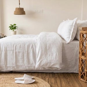 May include: A bed with white bedding, including a duvet and pillows, is the focal point of the image. A woven side table with a lamp and a woven rug are also visible. The room has a neutral color palette with natural textures.
