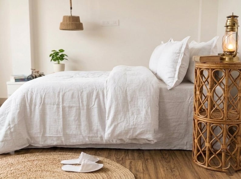 May include: A bed with white bedding, including a duvet and pillows, is the focal point of the image. A woven side table with a lamp and a woven rug are also visible. The room has a neutral color palette with natural textures.