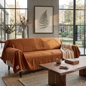 May include: A rust-colored linen sofa cover draped over a couch, featuring a small rectangular fabric label on the lower corner. The cover has a textured, natural weave and hangs loosely over the arms and seat, providing full coverage.