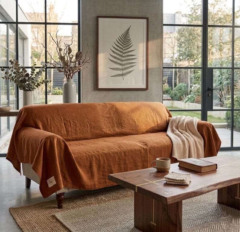 May include: A rust-colored linen sofa cover draped over a couch, featuring a small rectangular fabric label on the lower corner. The cover has a textured, natural weave and hangs loosely over the arms and seat, providing full coverage.