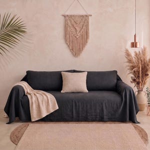 Könnte beinhalten: Ein dunkel anthrazitfarbenes Sofa mit strukturiertem Stoffbezug, in dessen Mitte ein quadratisches, hellbeiges Zierkissen liegt und über dessen linke Armlehne eine gefaltete, cremefarbene Strickdecke drapiert ist, vor einer zartrosa Wand.