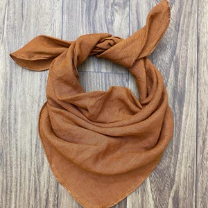 Pode incluir: Um bandana cor de ferrugem feito de um tecido macio e texturizado. O bandana está amarrado em um nó no topo e as pontas estão penduradas.