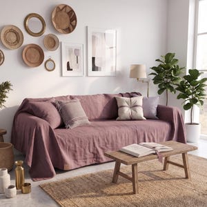 Op de afbeelding: Een mauve linnen bankhoes gedrapeerd over een bank, met een textuurrijke stoffen afwerking. De bank is geaccentueerd met verschillende sierkussens in neutrale tinten, waaronder een vierkant kussen met franjes en een effen rechthoekig kussen voor een gelaagde uitstraling.