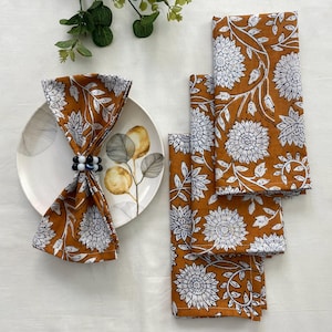 Op de afbeelding: Set van vier oranje en witte katoenen servetten met bloemenprint. De servetten zijn gevouwen en gestapeld op een wit oppervlak. De servetten hebben een witte bloemenprint op een oranje achtergrond.