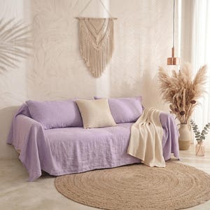 Puede incluir: Un sofá de color lavanda cubierto con una manta del mismo tono, que incluye dos cojines grandes de color lavanda y un cojín cuadrado de color crema. Una manta de punto de color crema está colocada sobre el lado derecho del sofá.