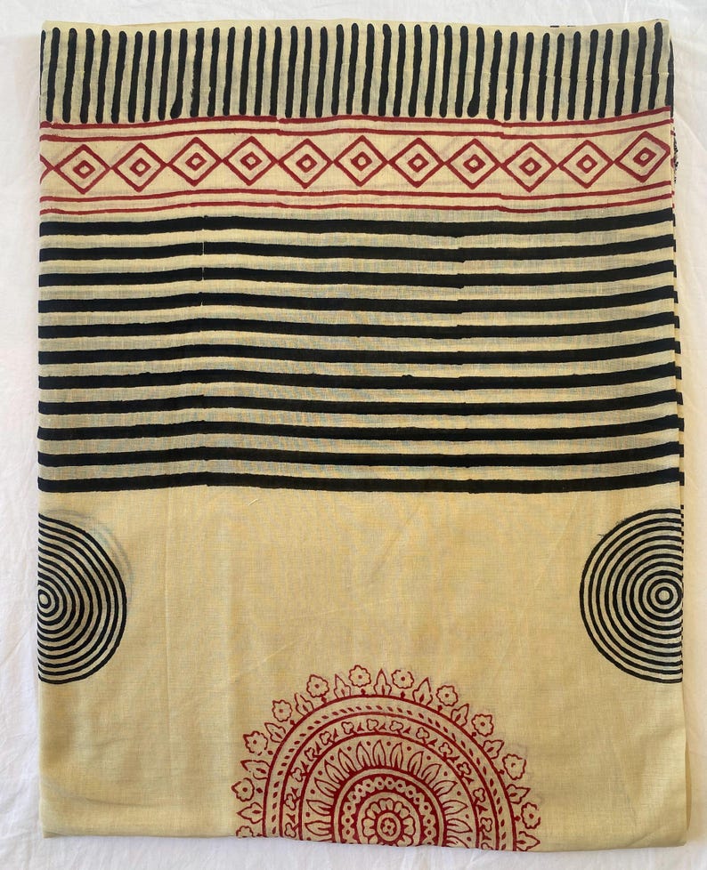 May include: Cream-colored fabric with a black and red geometric pattern. The design includes black stripes, vertical lines, and circular and diamond shapes. The fabric is likely used for textiles or home decor.