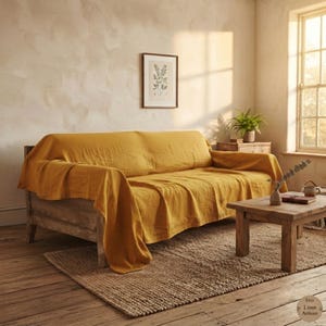 Könnte beinhalten: Eine senfgelbe Leinen-Überwurfdecke, die über ein Holzsofa drapiert ist. Der Stoff hat eine sichtbare, strukturierte Webart und hängt über die Armlehnen und die Sitzfläche des Möbels. Die Decke bedeckt die gesamte Sitzfläche und bildet weiche Falten.
