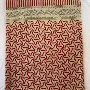 May include: A folded textile with a repeating red and cream-colored pattern. The design features a series of swirling, star-like shapes. The top edge has a striped border in red and a decorative band with cloud and triangle motifs.