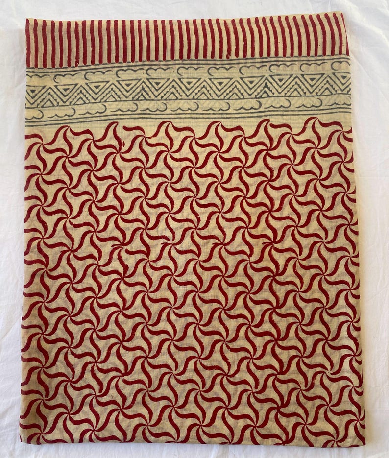 May include: A folded textile with a repeating red and cream-colored pattern. The design features a series of swirling, star-like shapes. The top edge has a striped border in red and a decorative band with cloud and triangle motifs.