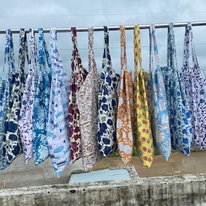 Pode incluir: Uma coleção de sacolas coloridas com vários designs florais e estampados. As sacolas são feitas de tecido e penduradas em uma barra de metal. As cores incluem azul, branco, amarelo, laranja e bordô.