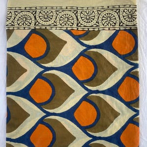 May include: A folded textile with a cream-colored top border featuring a black and cream floral pattern. The main body displays a repeating pattern of orange circles, blue teardrop shapes, and brown and cream elements.